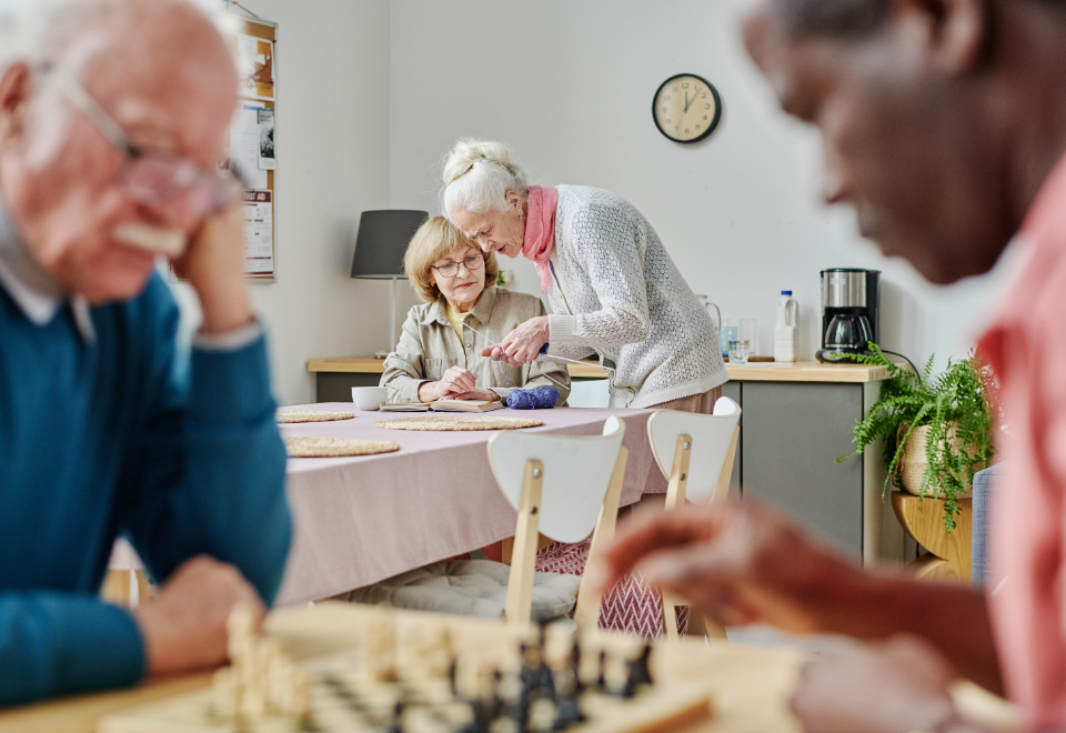 elderly men playing chess with elderly women knitting in the background